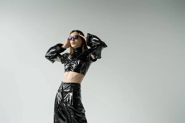 Stylish young woman in black latex outfit and sunglasses posing isolated on grey — Stock Photo