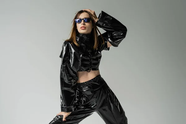 Stylish woman in black latex outfit and trendy sunglasses posing isolated on grey — Fotografia de Stock