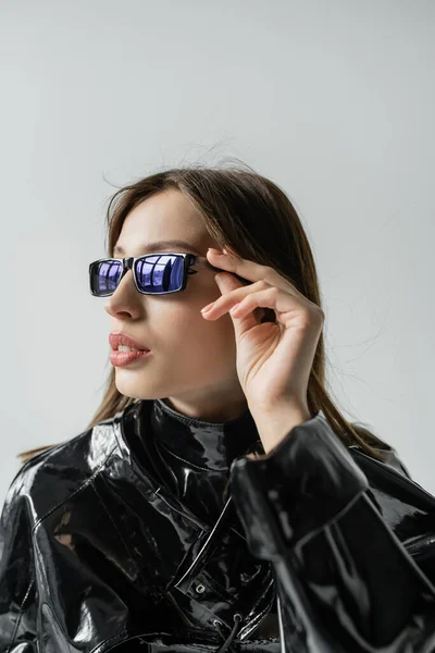 Young woman in black latex jacket adjusting trendy sunglasses isolated on grey — Foto stock