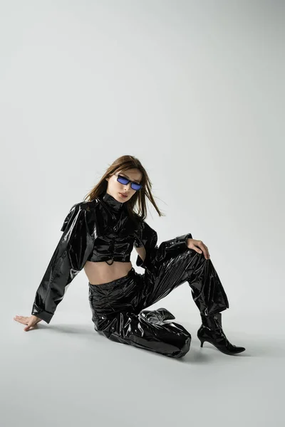 Full length of stylish young woman in black latex clothing and sunglasses sitting on grey — Photo de stock