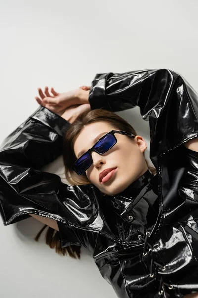 Top view of brunette woman in black latex jacket and stylish sunglasses lying on grey — Foto stock