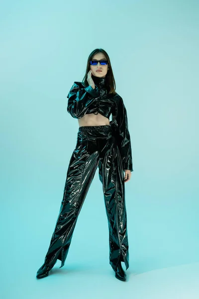 Full length of stylish young woman in black latex clothing and sunglasses standing on blue - foto de stock