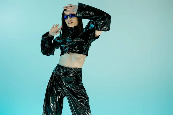 Stylish young woman in black latex clothing and sunglasses gesturing isolated on blue — Photo de stock