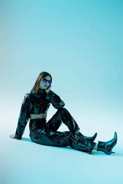 Full length of young woman in black latex clothing and high heels sitting on blue  background — Stock Photo