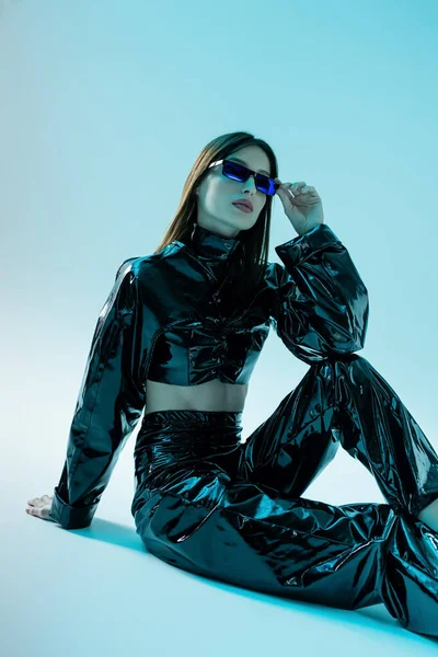 Young woman in black latex clothing adjusting sunglasses while sitting on blue — Foto stock