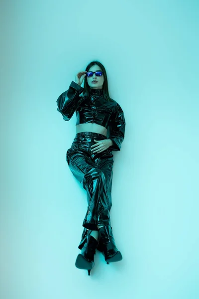 Full length of stylish young woman in black latex clothing and high heels adjusting sunglasses on blue - foto de stock