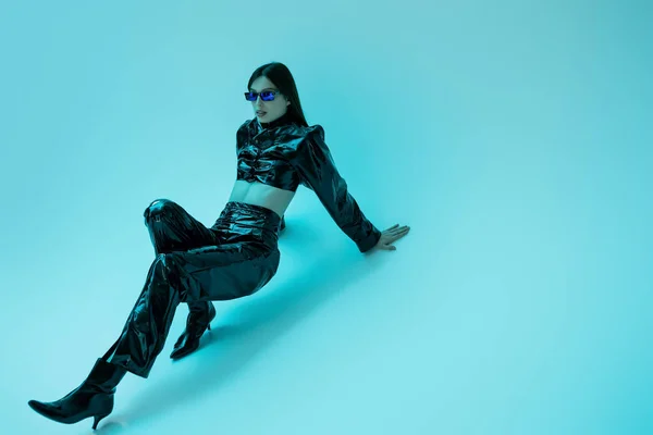 Full length of stylish young woman in latex clothing and high heels sitting on blue background — Stock Photo