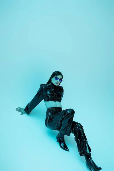 Full length of young woman in sexy latex clothing and high heels sitting on blue background — Photo de stock