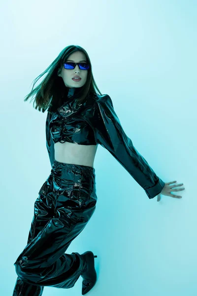 Brunette woman in sunglasses and sexy latex clothing posing on blue background — Stock Photo