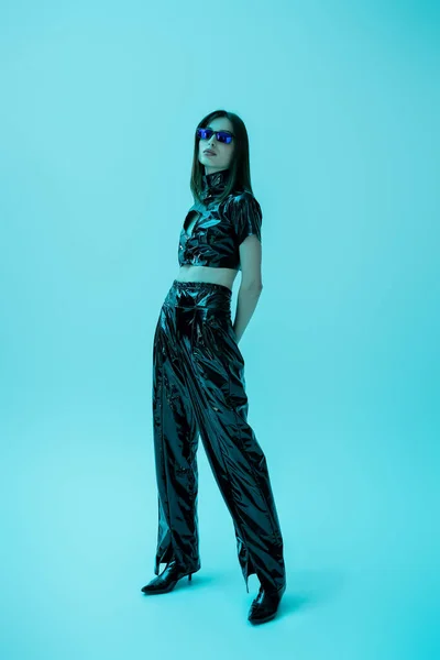 Full length of stylish young woman in latex crop top and trousers standing on blue — Stock Photo