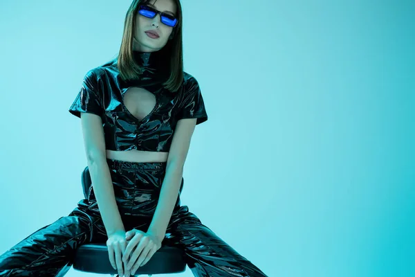 Stylish young woman in latex crop top and trousers sitting on stool while looking at camera isolated on blue — Stockfoto