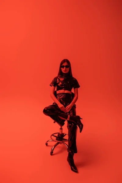 Full length of brunette woman in latex crop top and trousers sitting on stool while looking at camera on red — Stock Photo