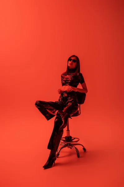 Full length of stylish woman in latex crop top and trousers sitting on stool while looking at camera on red - foto de stock