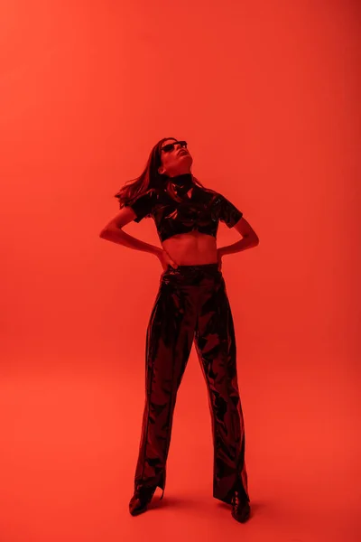 Full length of stylish woman in latex crop top and trousers standing with hands on hips on red — Stock Photo