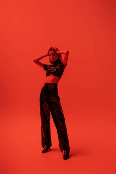 Full length of stylish woman in latex crop top and trousers posing with hands on head on red - foto de stock