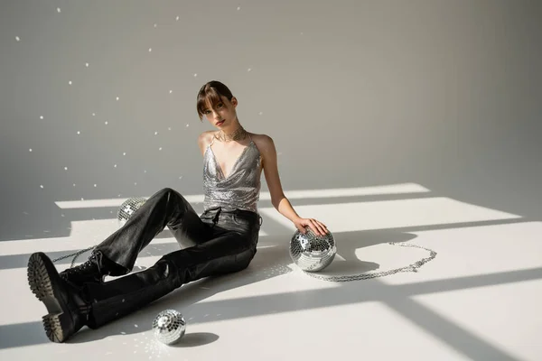 Full length of stylish woman in shiny top and leather pants sitting near disco balls on grey — Fotografia de Stock
