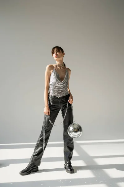 Повна довжина стильної жінки в блискучих верхніх і шкіряних штанях, що тримають ланцюг з диско-кулькою на сірому — стокове фото