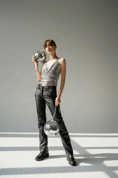 Full length of young woman in shiny top and leather pants holding chain with disco balls on grey — Stock Photo