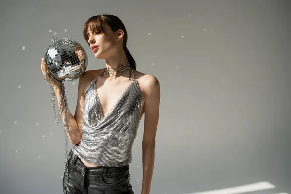 Young woman in trendy top and leather pants holding chain with disco ball on grey - foto de stock
