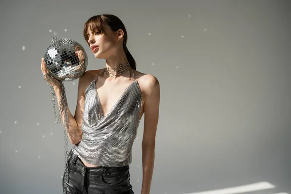 Young woman in trendy top and leather pants holding chain with disco ball on grey — Stockfoto