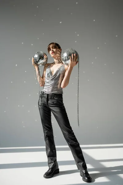 Full length of stylish woman in trendy top and leather pants holding shiny disco balls on grey — Stock Photo