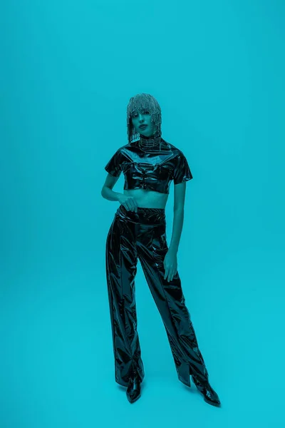 Full length of stylish model in headwear and latex clothes posing on blue background with lighting — Photo de stock