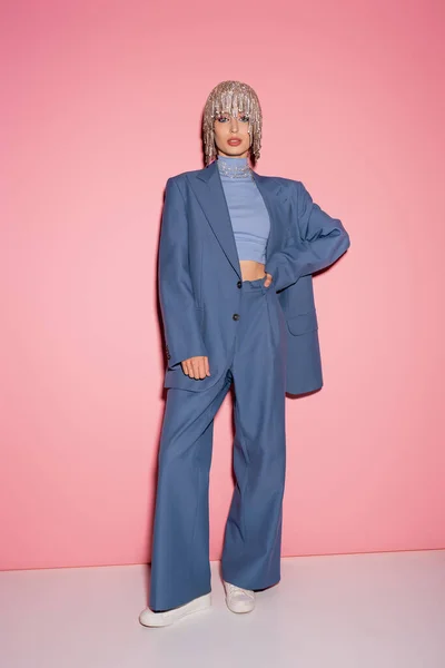 Full length of trendy woman in jewelry headwear and suit holding hand on hip on pink background — Photo de stock