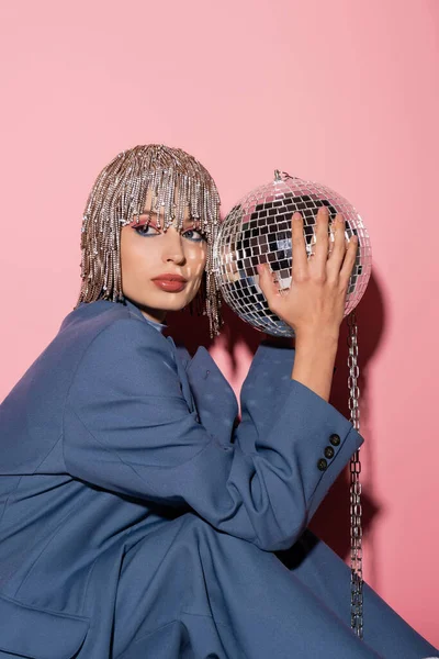 Stylish woman in jewelry headwear and jacket holding disco ball on pink background — Stockfoto