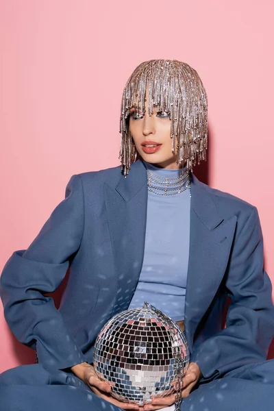 Fashionable young woman in blue suit and jewelry headwear holding disco ball on pink — Foto stock