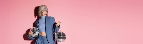 Trendy woman in luxury headwear and suit holding mirror balls on pink background, banner — Stock Photo
