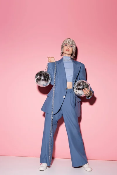 Full length of stylish model in blue suit and jewelry headwear holding disco balls on pink background — Stock Photo