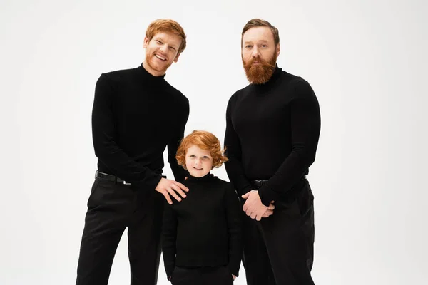 Redhead boy standing with hands in pockets near happy dad and grandpa while smiling at camera isolated on grey — Stock Photo
