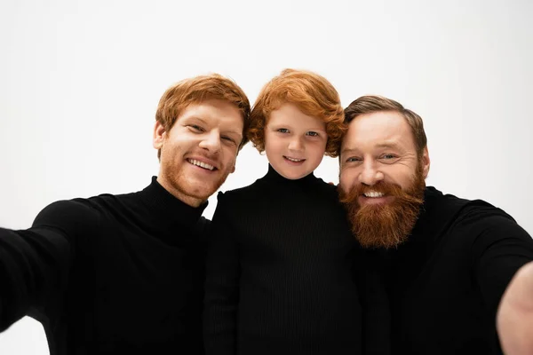 Joyful bearded men in black pullovers smiling at camera near red haired boy isolated on grey — Stock Photo