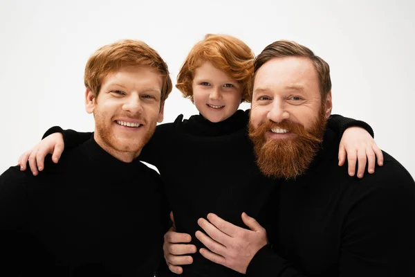 Smiling redhead kid hugging bearded grandpa and dad wearing black turtlenecks isolated on grey — Stock Photo