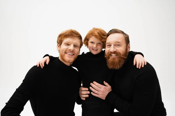 Joyful red haired kid embracing dad and bearded grandfather in black sweaters isolated on grey — Foto stock