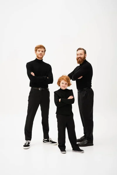 Full length of red haired kid with father and bearded granddad in black clothes standing with crossed arms on grey background — Stock Photo