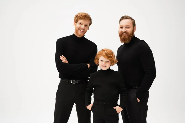 Joyful redhead kid standing with hands in pockets near bearded dad and grandfather isolated on grey — Photo de stock