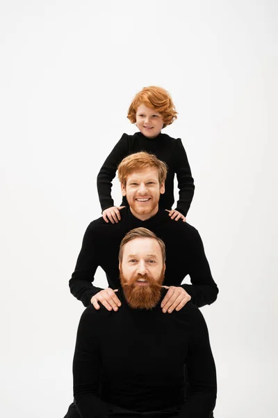Cheerful red haired kid posing near bearded dad and grandfather in black turtlenecks isolated on grey — Foto stock