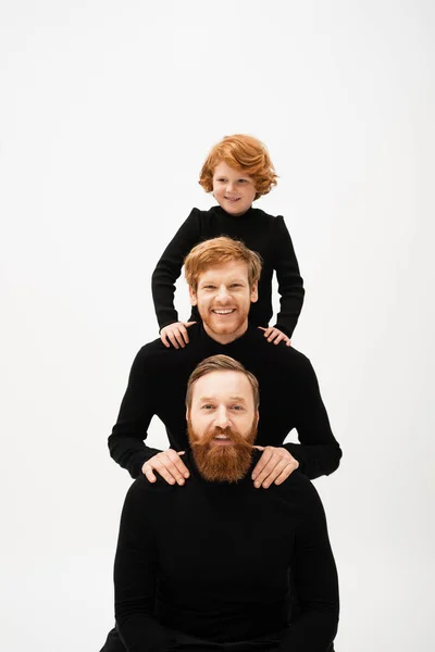 Cheerful red haired kid posing near bearded dad and grandfather in black turtlenecks isolated on grey — Stock Photo