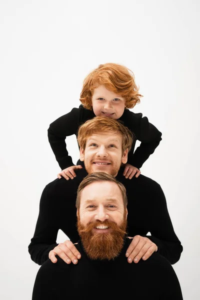 Joyful redhead kid posing with smiling and bearded grandpa and dad in black turtlenecks isolated on grey — Stockfoto