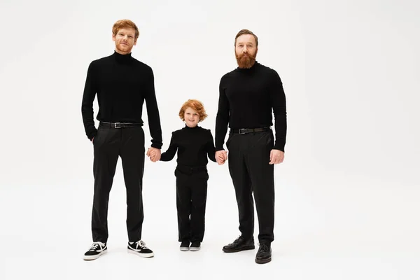Full length of bearded men in black clothes holding hands with joyful redhead boy on light grey background — Photo de stock