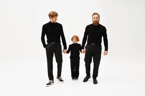 Full length of bearded men in black turtlenecks and trousers holding hands with happy red haired kid on light grey background — Stock Photo