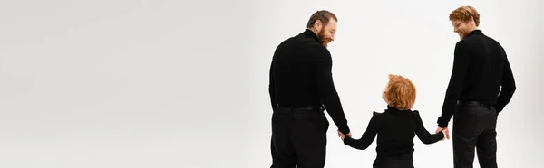 Back view of redhead boy holding hands with smiling father and bearded granddad in black clothes isolated on grey, banner — Stock Photo