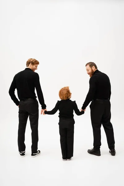 Full length of bearded men in black clothes smiling at redhead boy while holding hands on light grey background — Stock Photo