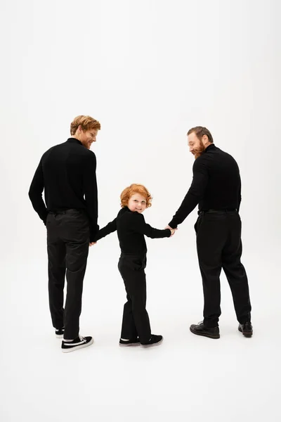 Full length of cheerful redhead boy looking at camera while holding hands with dad and grandpa in black clothes on light grey background — Stockfoto
