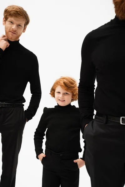 Cheerful kid in black clothes looking at camera while posing with hands in pockets near redhead father and grandpa isolated on grey — Stock Photo