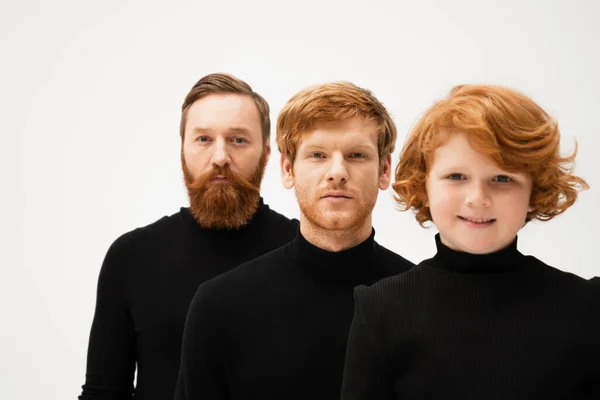 Happy red haired boy smiling at camera near father and bearded granddad in black sweaters isolated on grey — Stock Photo