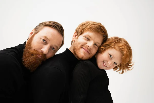 Cheerful redhead kid smiling at camera near bearded grandpa and dad leaning on each other isolated on grey — Foto stock