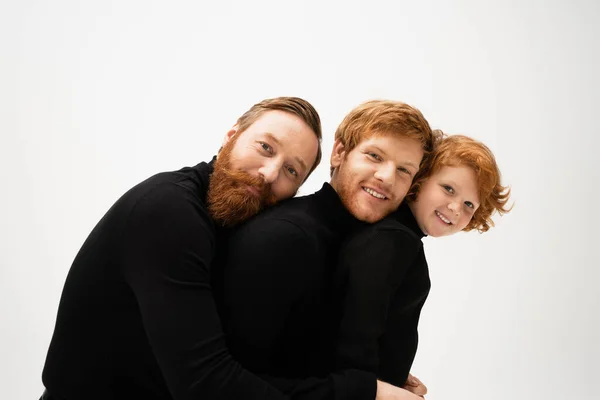 Cheerful redhead men and kid in black sweaters leaning on each other and smiling at camera isolated on grey — Foto stock