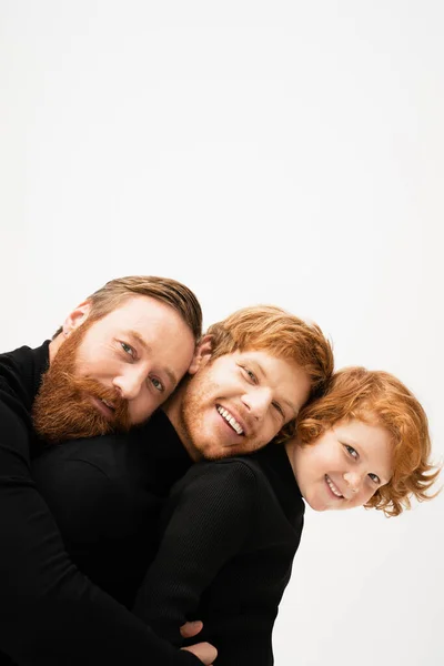 Joyful bearded men and redhead kid in black pullovers leaning on each other and smiling at camera isolated on grey — Stock Photo
